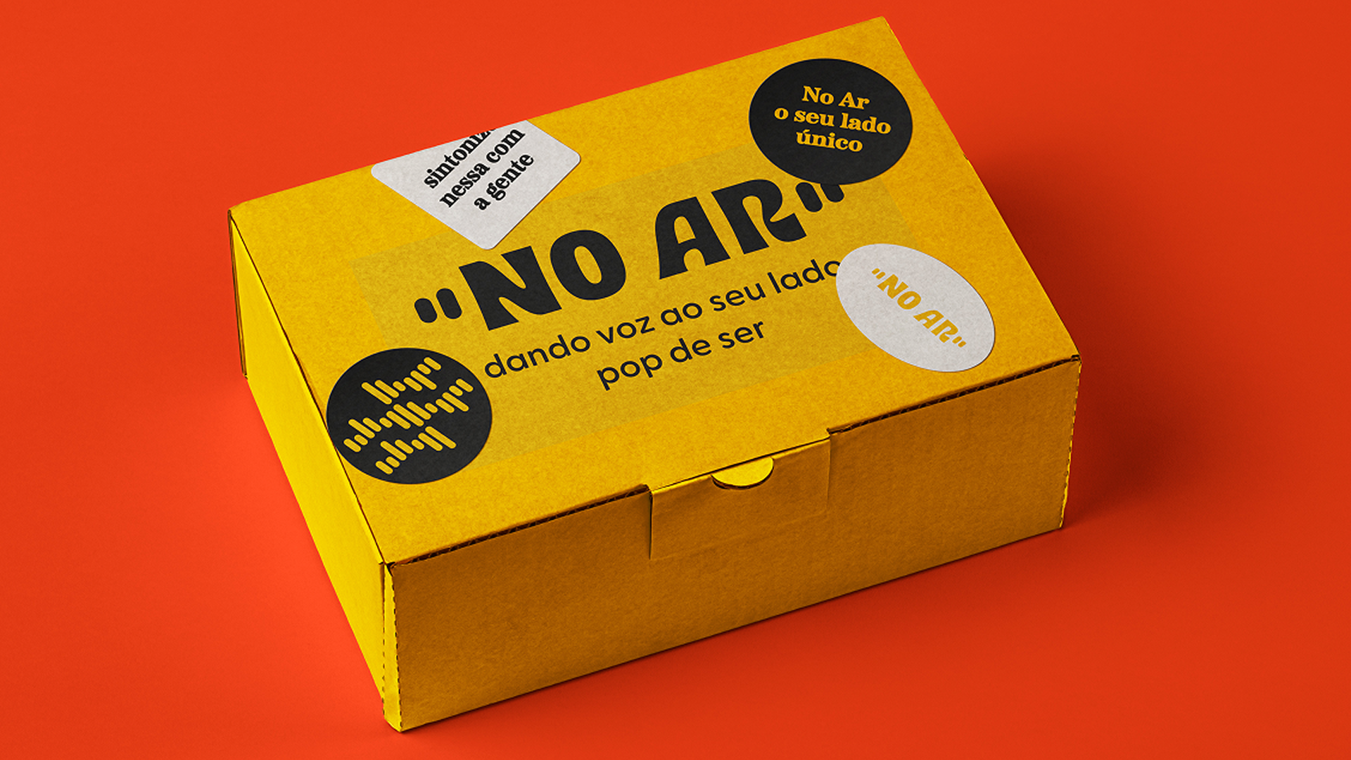 Caixa de envio do ecommerce criada para a marca No Ar na cor amarela com logo e adesivos pretos. A caixa também tem o slogan da marca "dando voz ao seu lado pop de ser".