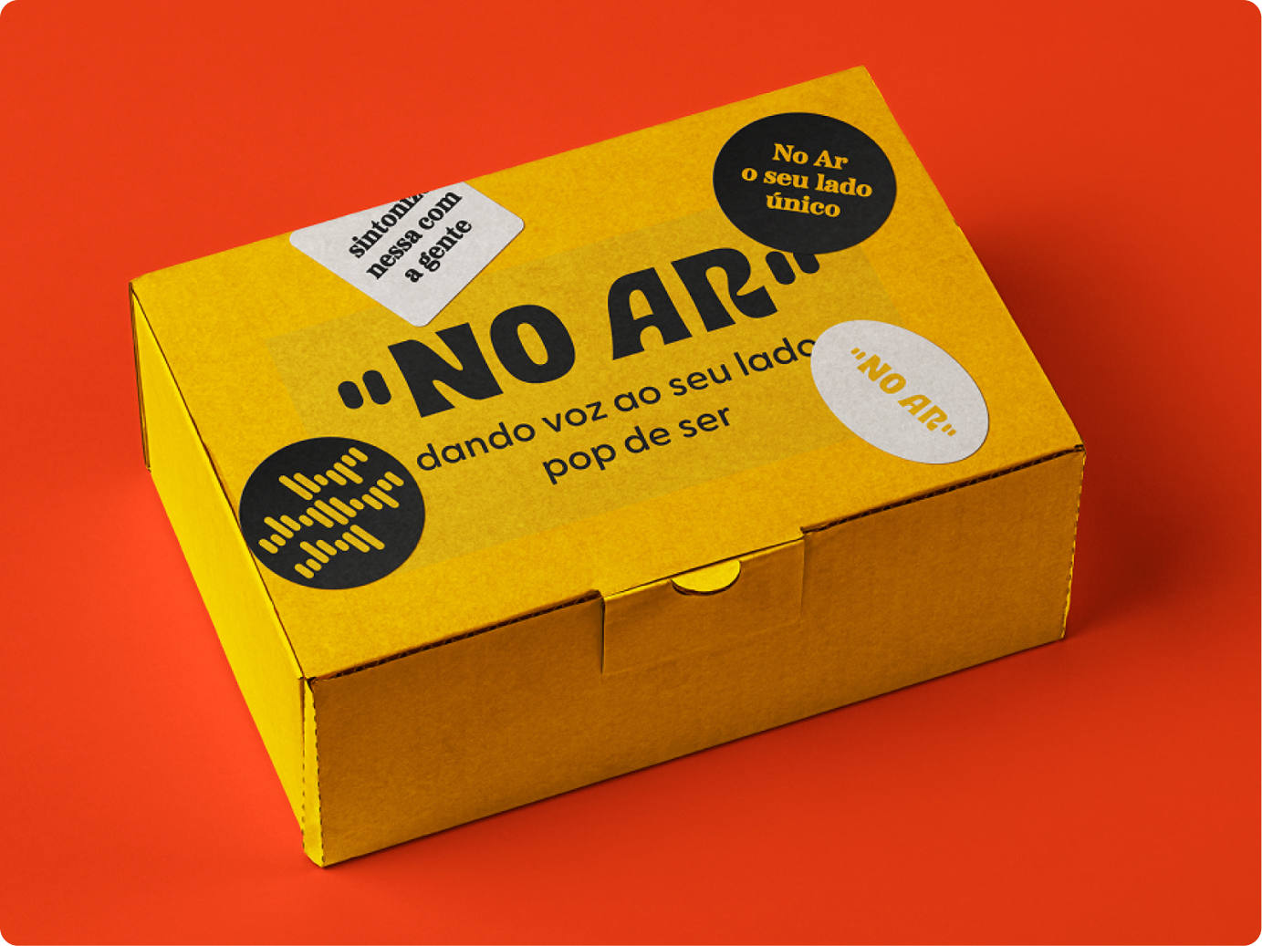 Embalagem de envios personalizada com a marca No Ar. A caixa é amarela com logo, slogan em preto e adesivos pregados.